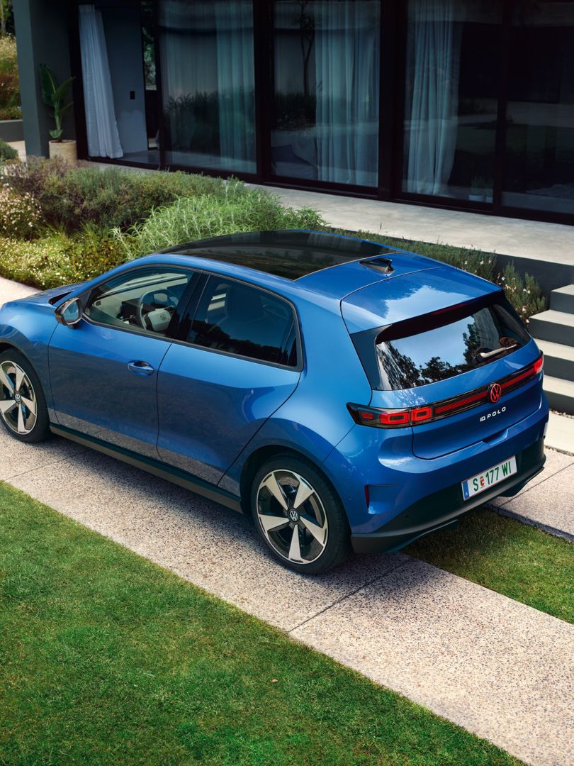 Modrý VW ID. Polo Style stojí zaparkovaný pred obytným domom so zeleňou, za vozidlom prechádza muž.