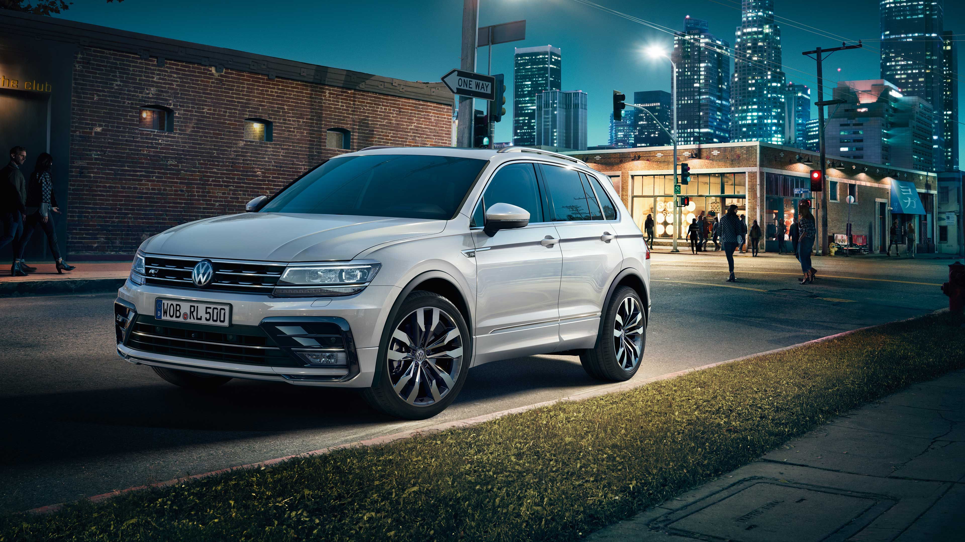 Biele SUV značky Volkswagen zaparkované na ceste na ulici za súmraku. V pozadí je osvetlené mesto, ľudia kráčajú po chodníku okolo moderných budov. Scéna vyvoláva mestskú atmosféru.