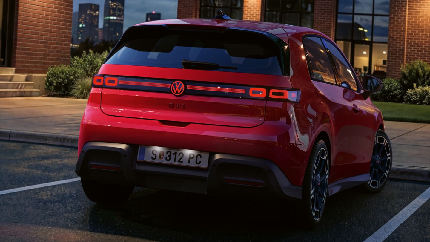 Pohľad na zadnú časť červeného VW ID. Polo GTI so zapnutými zadnými svetlami a osvetleným logom, ktoré parkuje pred administratívnou budovou.