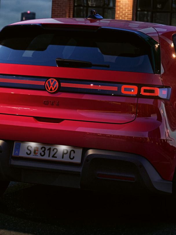 Pohľad na zadnú časť červeného VW ID. Polo GTI so zapnutými zadnými svetlami a osvetleným logom, ktoré parkuje pred administratívnou budovou.