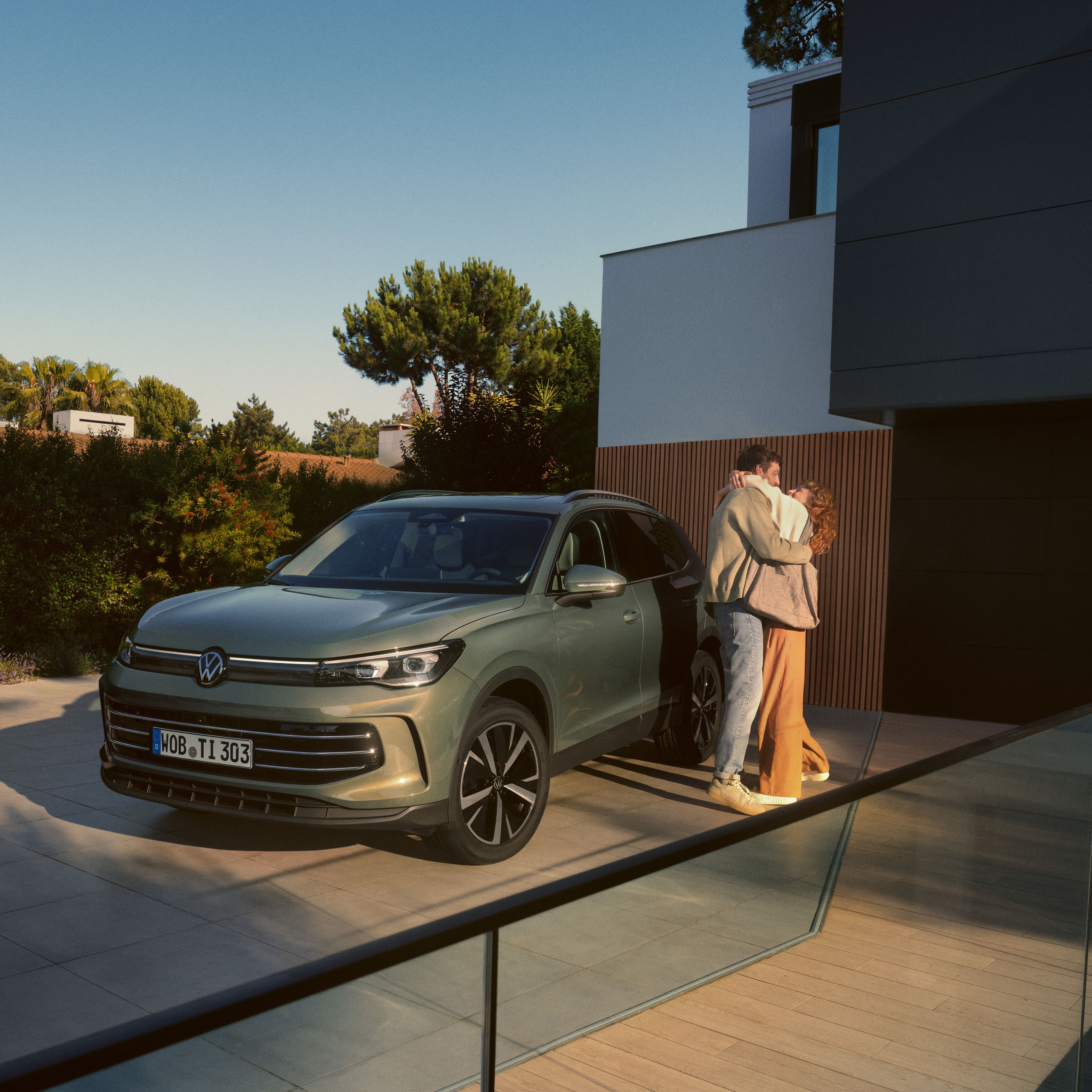 Zelený VW Tiguan zaparkovaný pre moderným domom.dernen Wohnhauses. Ein junges Paar steht daneben und umarmt sich.