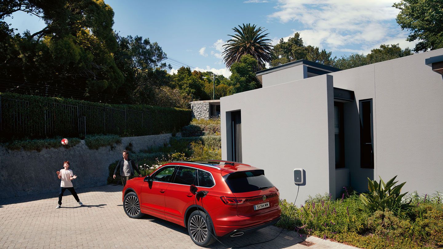 VW Touareg Elegance eHybrid, pohľad šikmo zozadu, sa nabíja pred moderným domom.