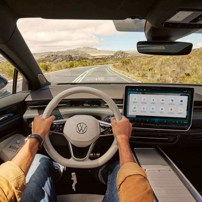 Muž sedí za volantom VW ID.7 s head-up displejom s rozšírenou realitou. Vedľa displeja je veľký displej systému infotainment. Na displeji sú ikony funkcií a aplikácií. Cez čelné sklo vidieť cestu v horách a zeleň, ktorá ju lemuje. V pozadí vidieť kopce.