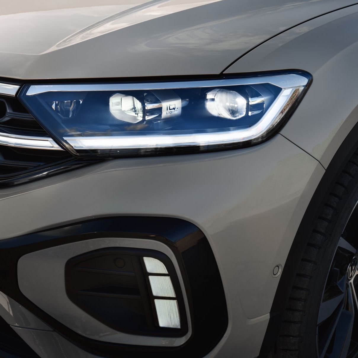 Detailný záber na predný svetlomet IQ.Light šedého SUV T-Roc. Svetlo je zasvietené. Vidieť tiež časť kapoty vozidla, predného nárazníka a masky chladiča. Čiastočne vidieť ak predné koleso s moderným diskom.