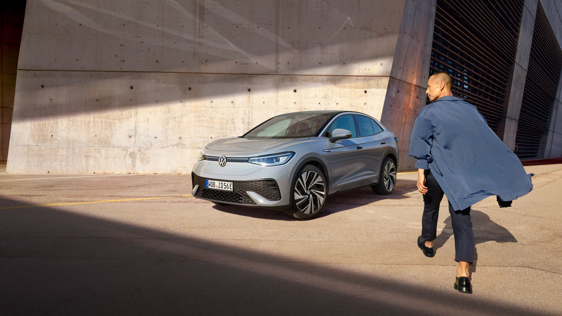 Moderné elektrické vozidlo Volkswagen ID.5 stojí vedľa betónovej steny na slnkom osvetlenej ulici. Muž s modrým kabátom kráča smerom k autu.