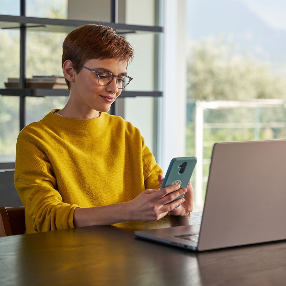 Žena sa pozerá na svoj mobil a sedí pred notebookom.
