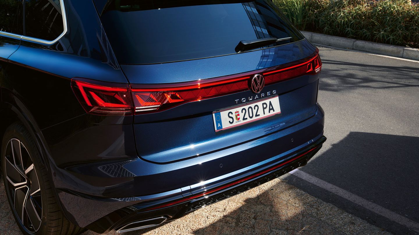 Pohľad na zadné partie modrého Volkswagen Touareg. Auto je zaparkované na parkovacom mieste pri ceste v meste. V pozadí vidieť pás zelene a chodník.
