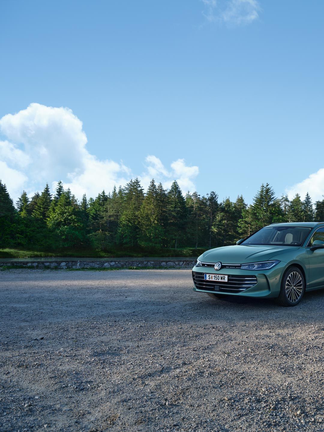 Zelený VW Passat je zaparkovaný na štrkovom priestore v lese. Atmosféru dotvára modrá obloha. Automobil je typu kombi. Scéna je pokojná a slnečná.
