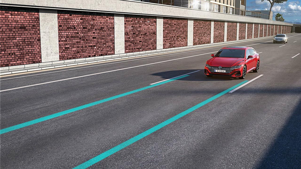 Lane assist a Side assist zobrazené ako dve modré línie jedného jazdného pruhu, v ktorom jazdí červené vozidlo. Vo vedľajšom pruhu ide druhé strieborné vozidlo. V pozadí vidieť oporný múr a modernú stavbu a stromy.