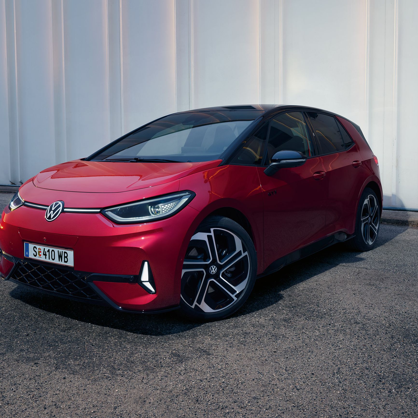 Červený elektromobil Volkswagen ID.3 parkuje na ceste pred bielou stenou. Vozidlo má moderný dizajn a čiernu sklenenú strechu.
