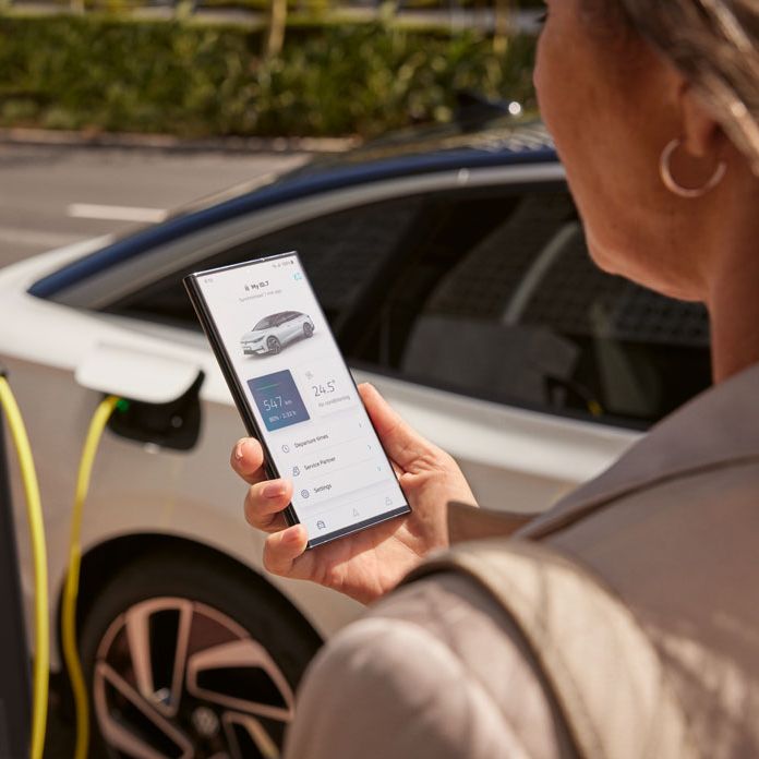 Žena stojí pri nabíjacej stanici pre elektromobily a drží telefón, na ktorom sleduje aplikáciu pre správu nabíjania auta. Nabíjačka je pripojená k bielemu elektromobilu. V pozadí je ulica a zeleň.