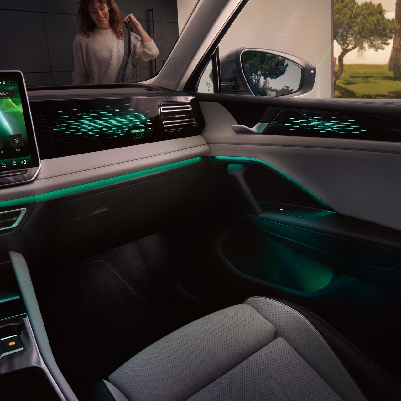 Pohľad do interiéru modeli Tiguan zo sedadla vodiča na miesto spolujazdca, čiastočne vidieť elegantnú palubnú dosku s ambientným osvetlením, centrálny displej systému infotainment, obklad dverí. Cez okná vidieť ženu prichádzajúcu k vozidlu od rodinného domu.