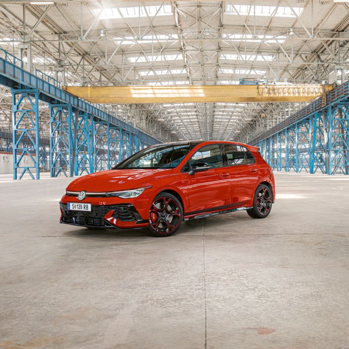 Červený Golf GTI je zaparkovaný vo veľkej, prázdnej priemyselnej budove s modrými oceľovými stĺpmi a nosníkmi. Strecha budovy je zo skla a prepúšťa dovnútra dostatok svetla.