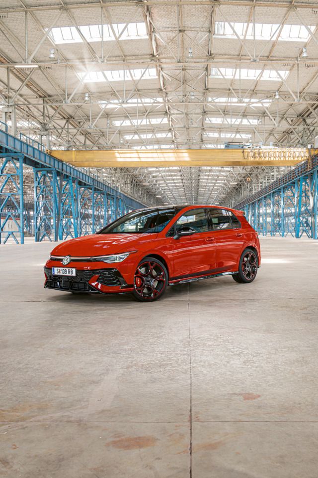 Červený Golf GTI je zaparkovaný vo veľkej, prázdnej priemyselnej budove s modrými oceľovými stĺpmi a nosníkmi. Strecha budovy je zo skla a prepúšťa dovnútra dostatok svetla.