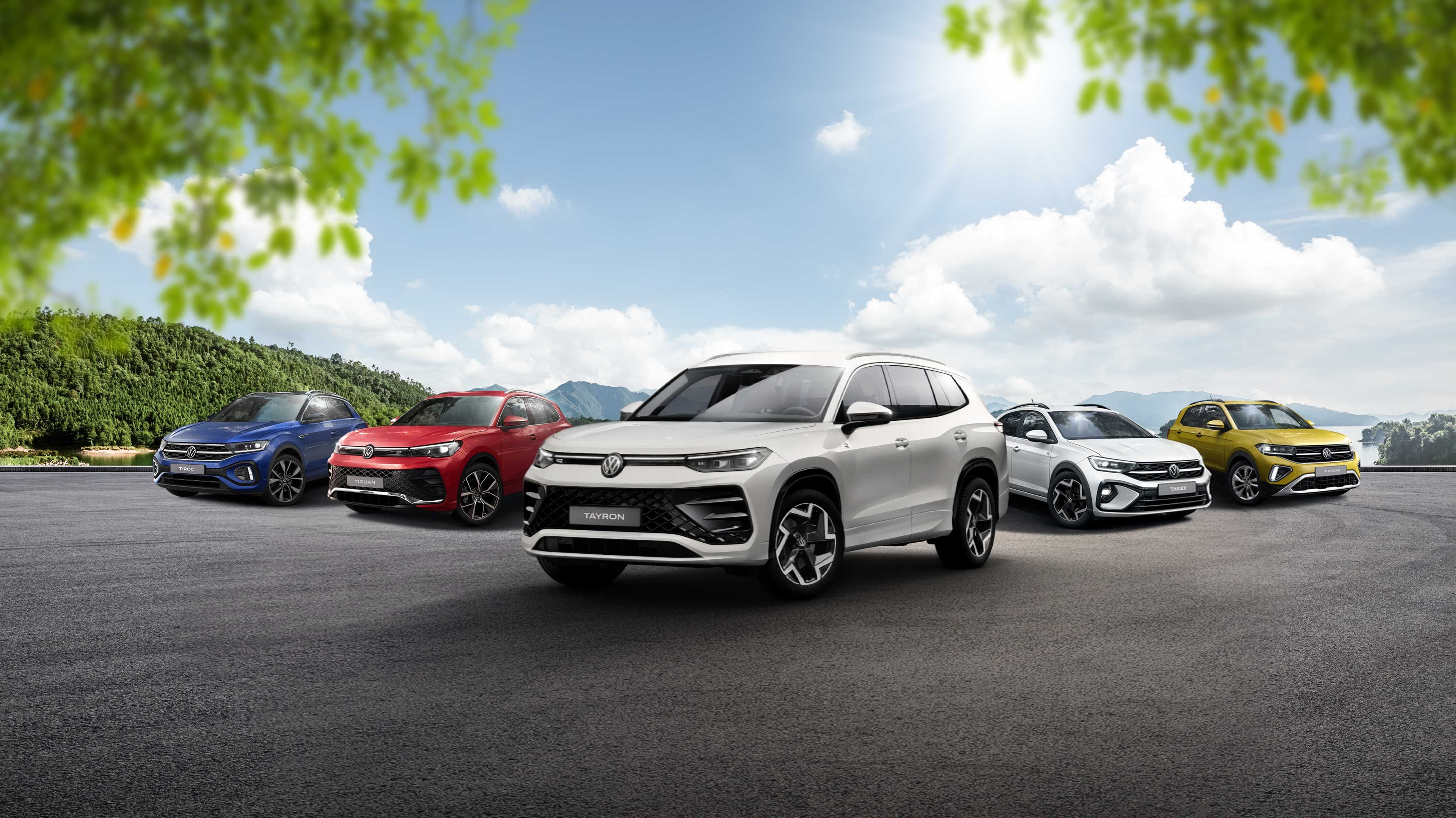 Päť rôznych vozidiel Volkswagen rôznych modelov a farieb zaparkovaných na otvorenom priestranstve. Automobil v popredí je veľké biele SUV. Okolie tvorí modrá obloha s oblakmi a stromy.