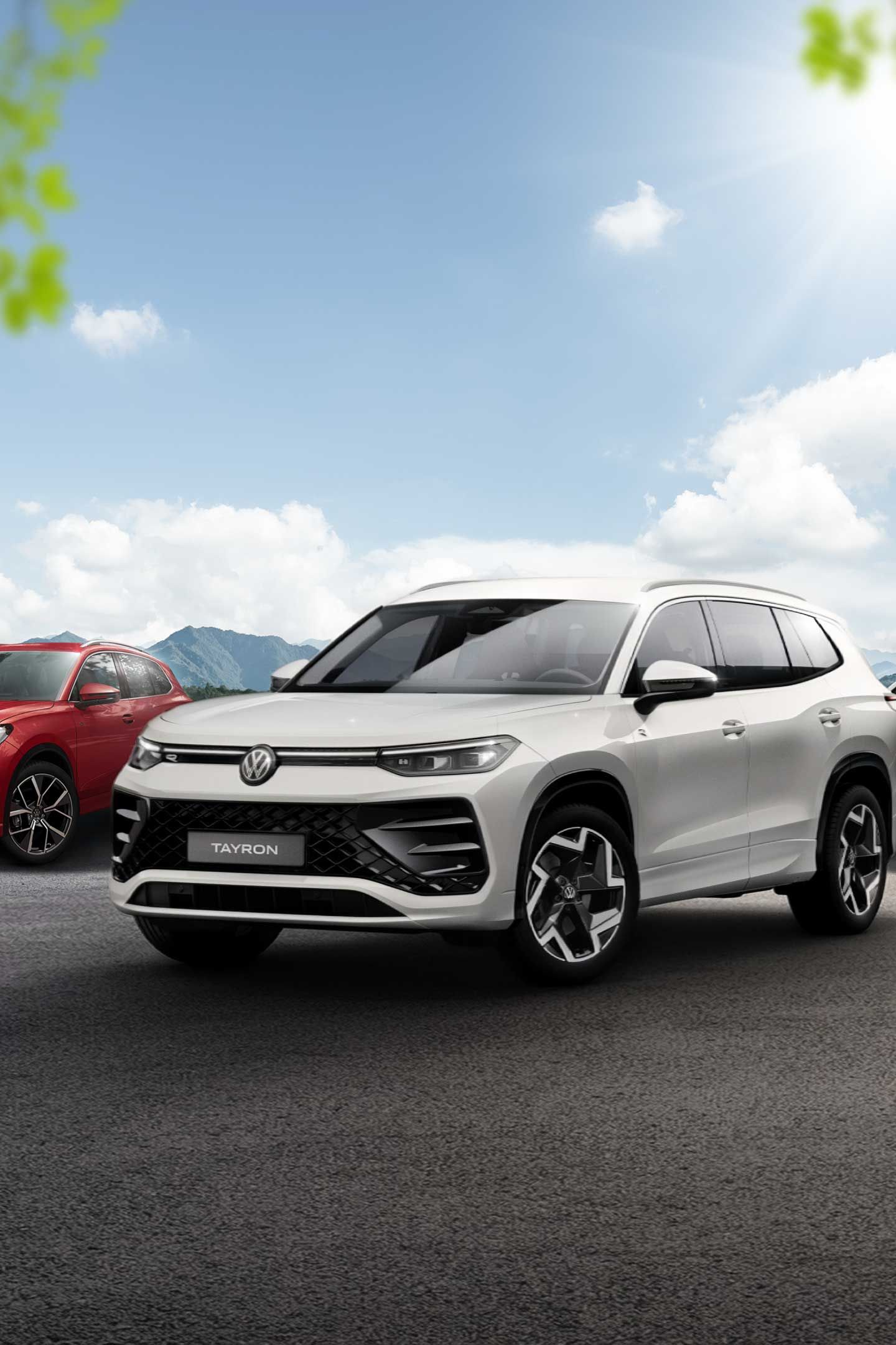 Päť rôznych vozidiel Volkswagen rôznych modelov a farieb zaparkovaných na otvorenom priestranstve. Automobil v popredí je veľké biele SUV. Okolie tvorí modrá obloha s oblakmi a stromy.