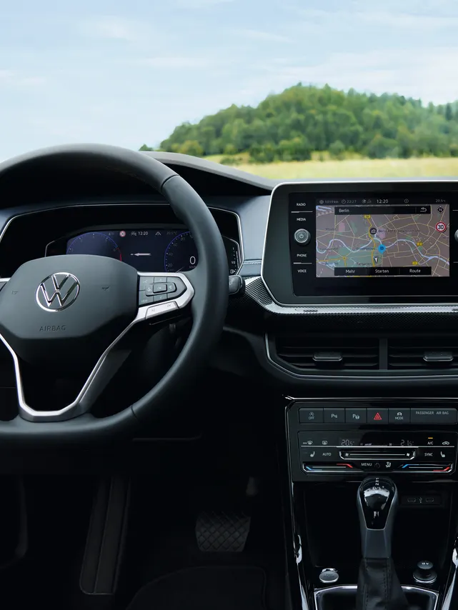 Detailný pohľad z miesta vodiča vo VW T-Cross na palubnú dosku, volant, prístrojový panel, displej systému infotainment. Cez čelné sklo vidieť lúku, kopec a lesy.