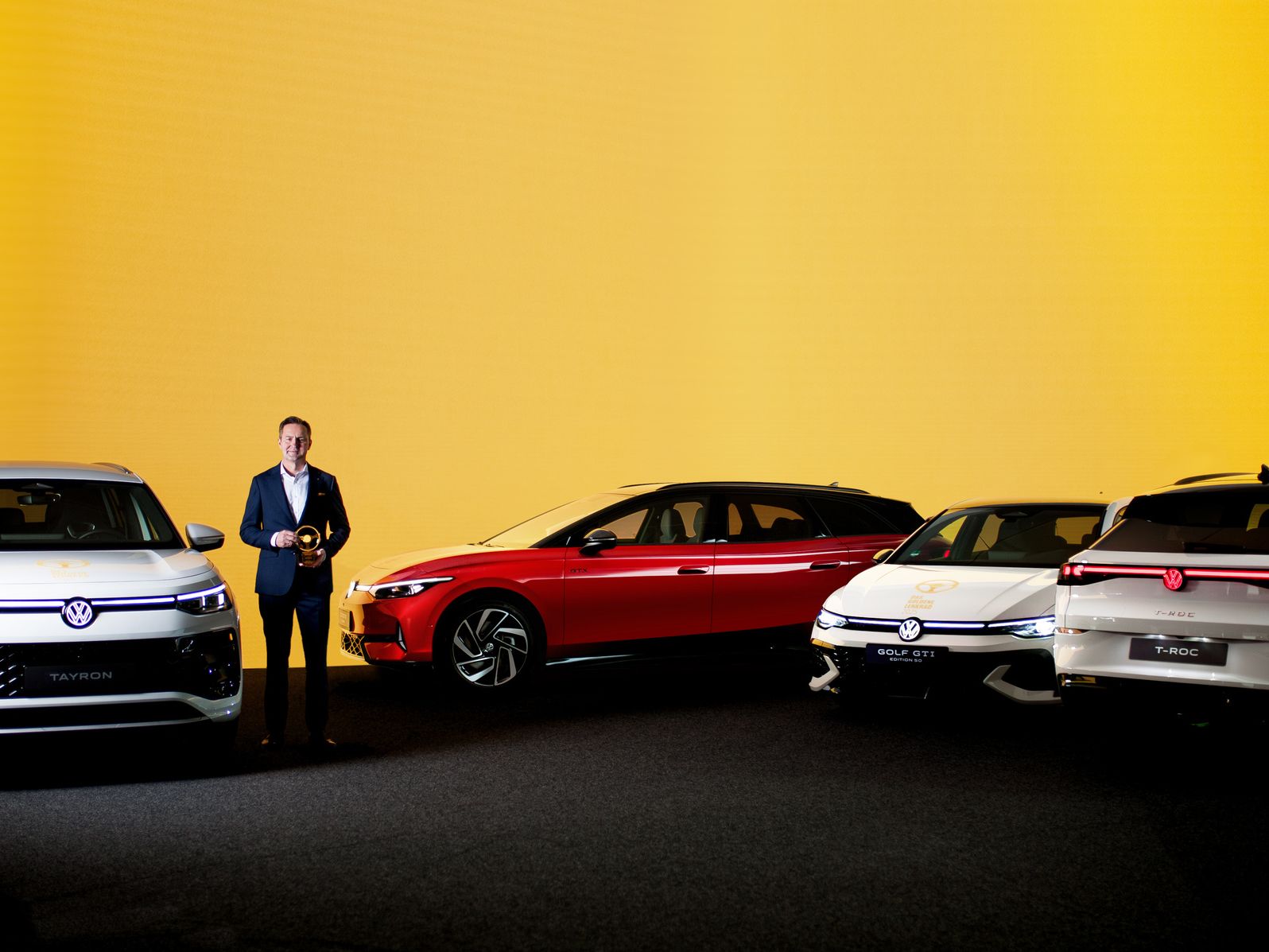 Exponát štyroch vozidiel značky Volkswagen, vystavených na pódiu pred svetlo žltým pozadím. Osoba stojí vedľa automobilov, držiac ocenenie.