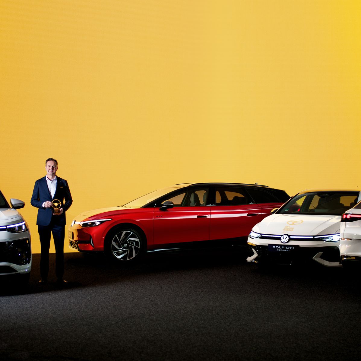 Exponát štyroch vozidiel značky Volkswagen, vystavených na pódiu pred svetlo žltým pozadím. Osoba stojí vedľa automobilov, držiac ocenenie.