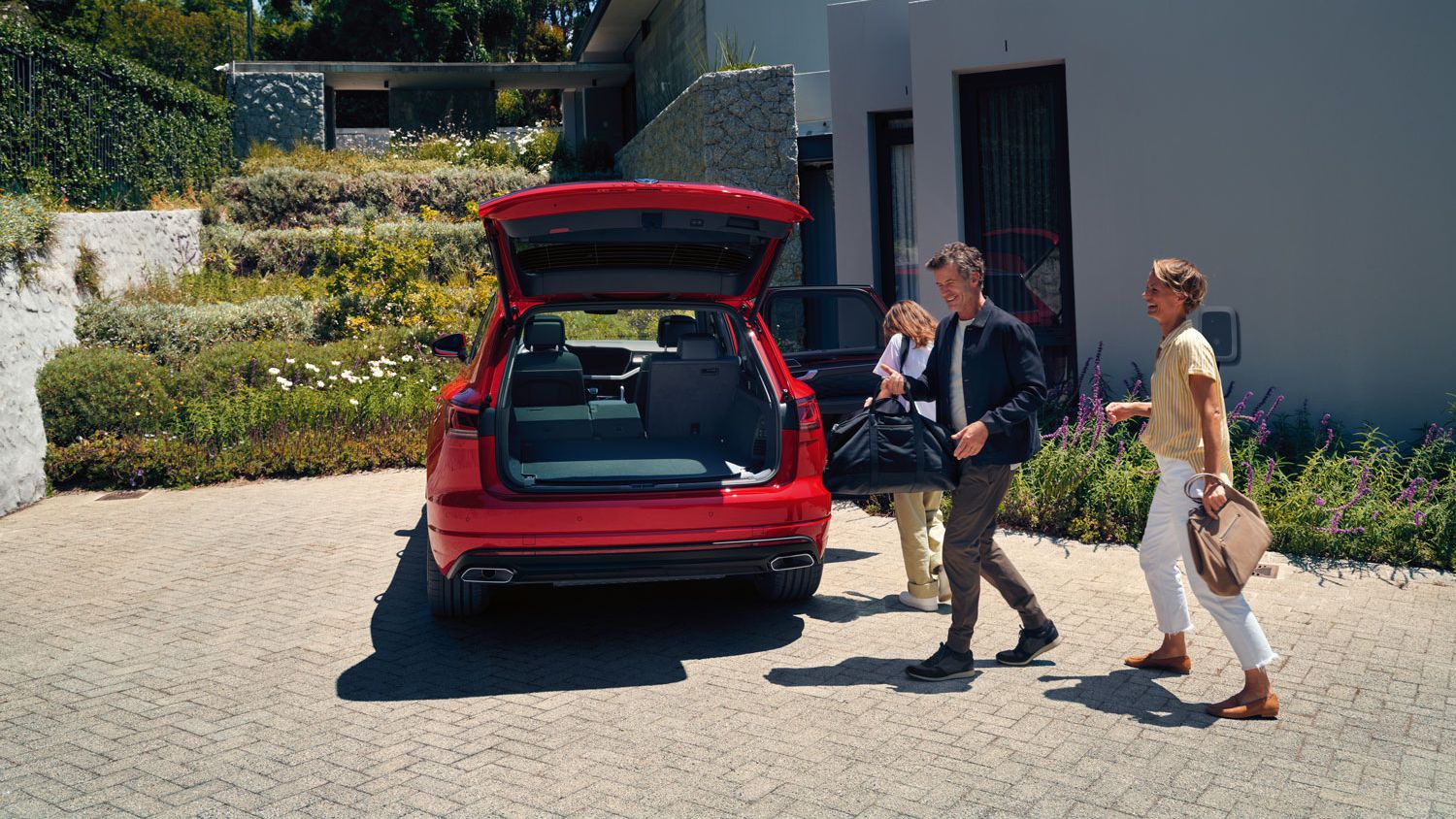 Muž, žena a dieťa idú k vozidlu VW Touareg Elegance eHybrid s otvoreným batožinovým priestorom. Vozidlo je zaparkované pri dome.