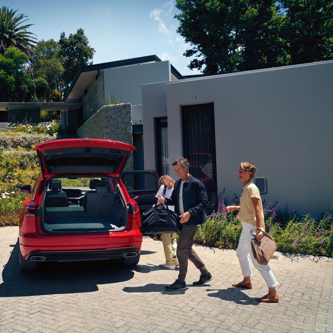 Muž, žena a dieťa idú k vozidlu VW Touareg Elegance eHybrid s otvoreným batožinovým priestorom. Vozidlo je zaparkované pri dome.