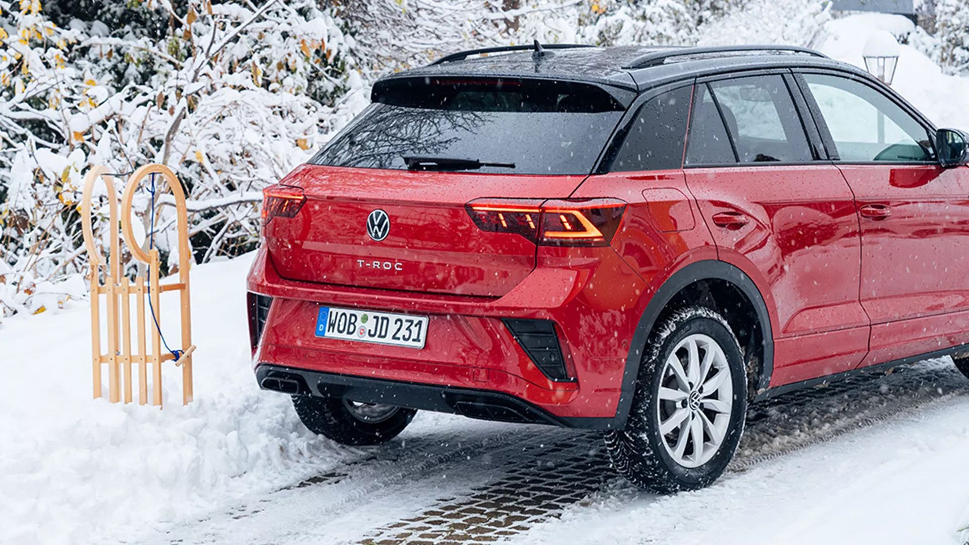 Červené auto Volkswagen T-Roc zaparkované na zasneženej ceste vedľa drevených saní. V pozadí sú zasnežené kríky. Cesta je pokrytá snehom.