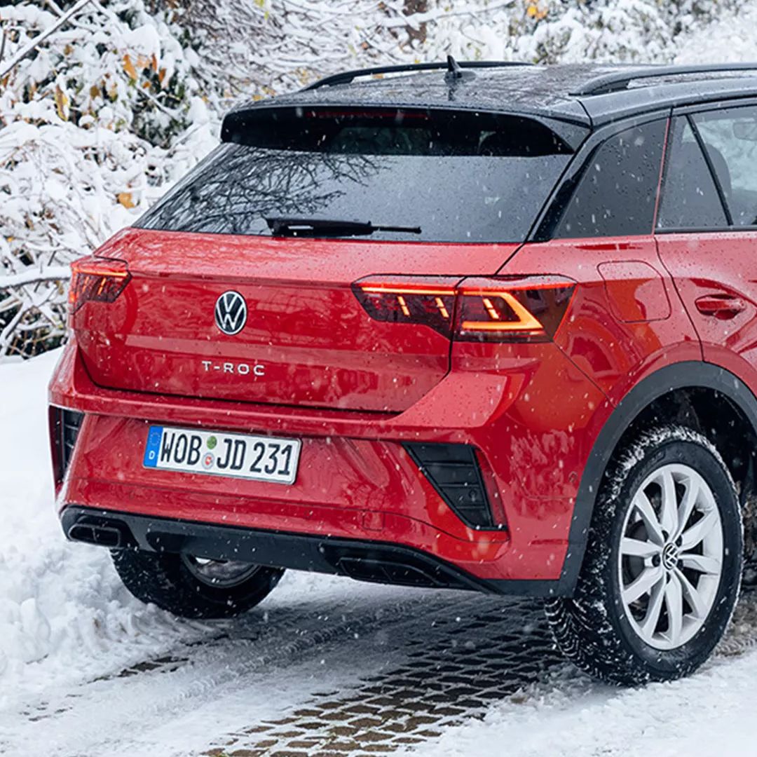 Červené auto Volkswagen T-Roc zaparkované na zasneženej ceste vedľa drevených saní. V pozadí sú zasnežené kríky. Cesta je pokrytá snehom.