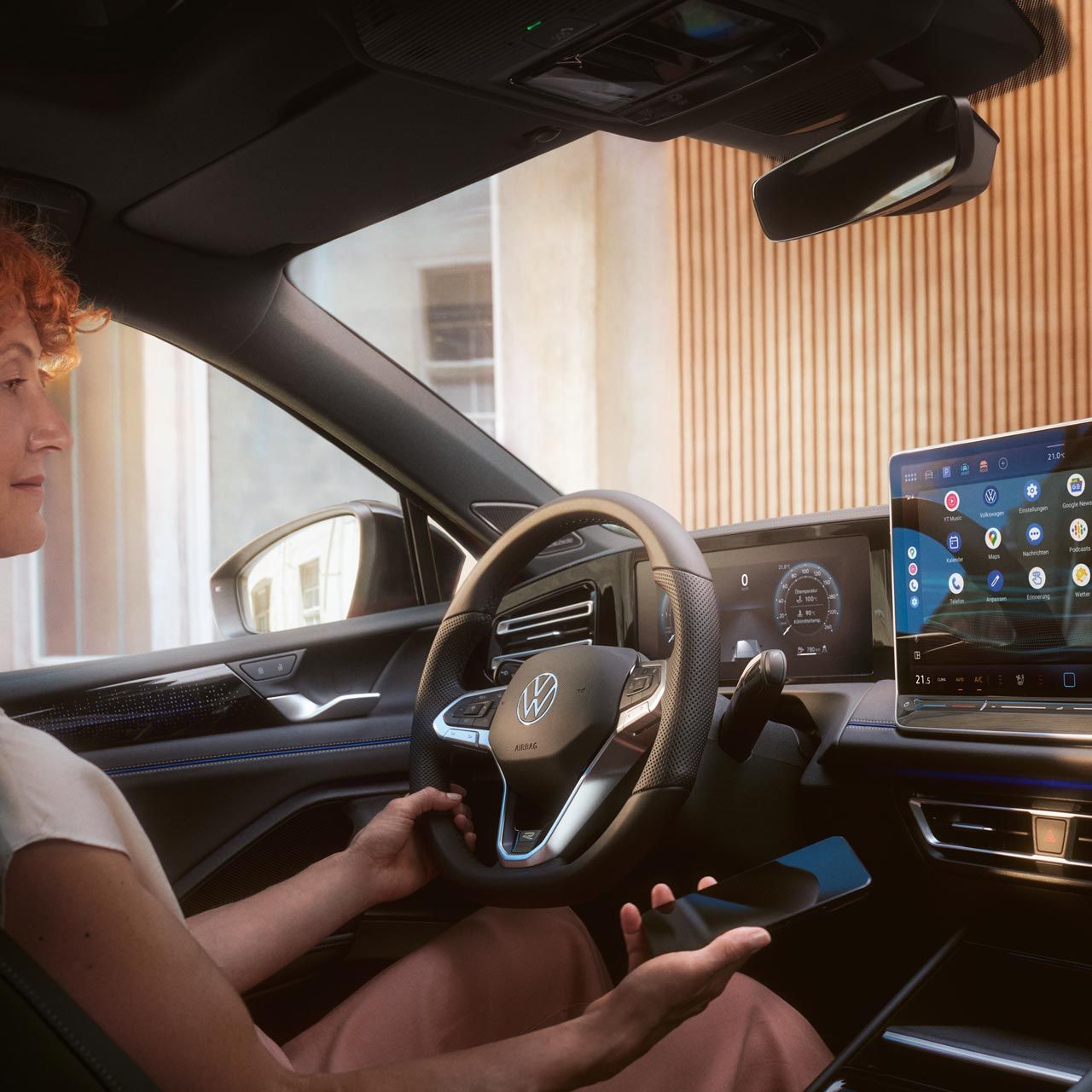 Žena sedí za volantom vozidla VW Tiguan. V ruke drží smartfón a pozerá sa na displej systému infotainment. Druhou rukou drží volant. Cez čelné sklo vidieť moderný dom.