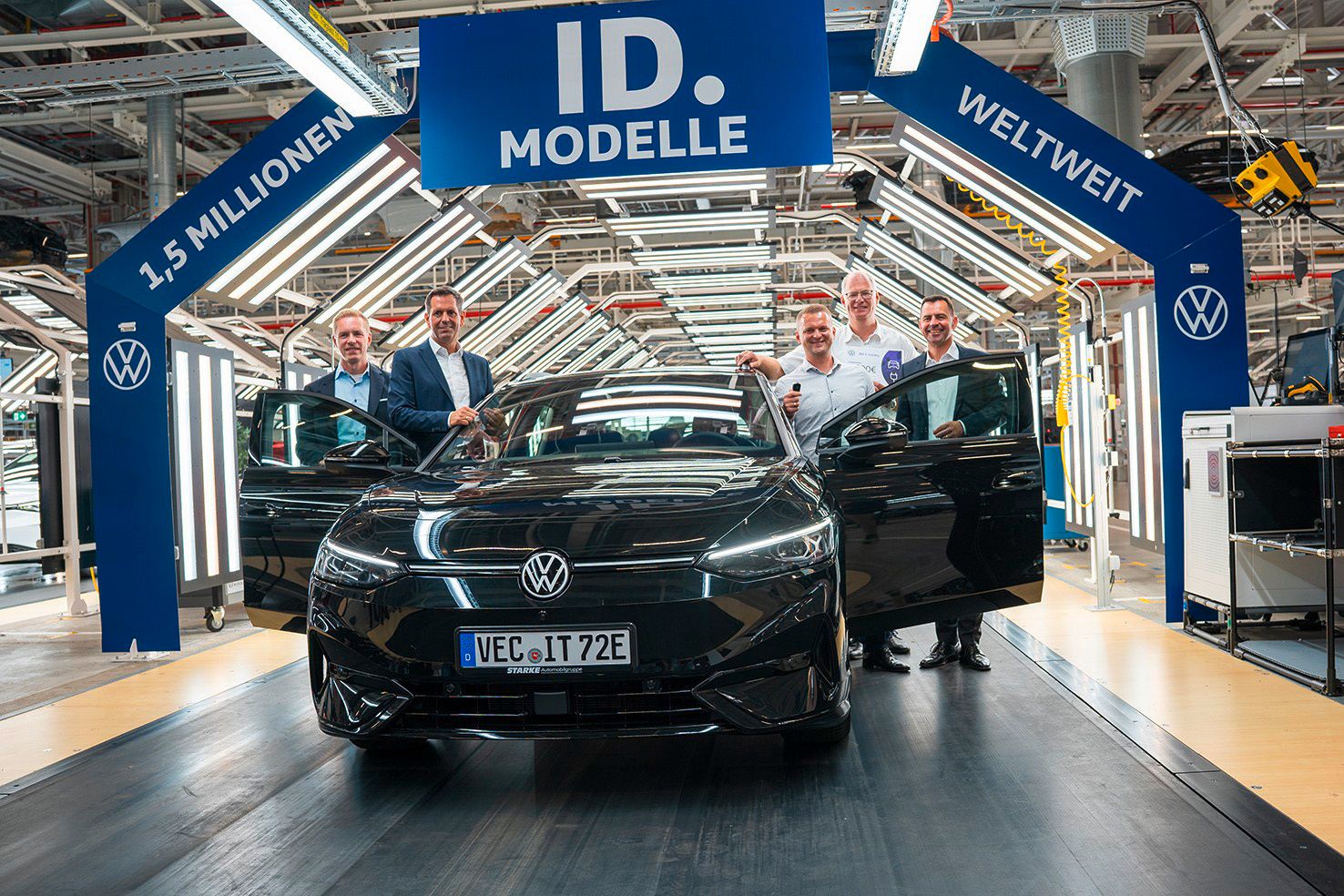 Automobil v továrni obklopený piatimi mužmi. Nad automobilom je veľký modrý banner s nápisom "1,5 Milionen ID. Modelle weltweit". V pozadí vidieť výrobné zariadenia.