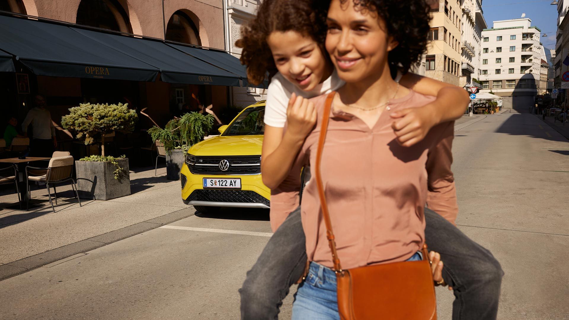 Na ulici v centre mesta nesie žena na chrbte dievča a žltý VW T-Cross stojí v pozadí.