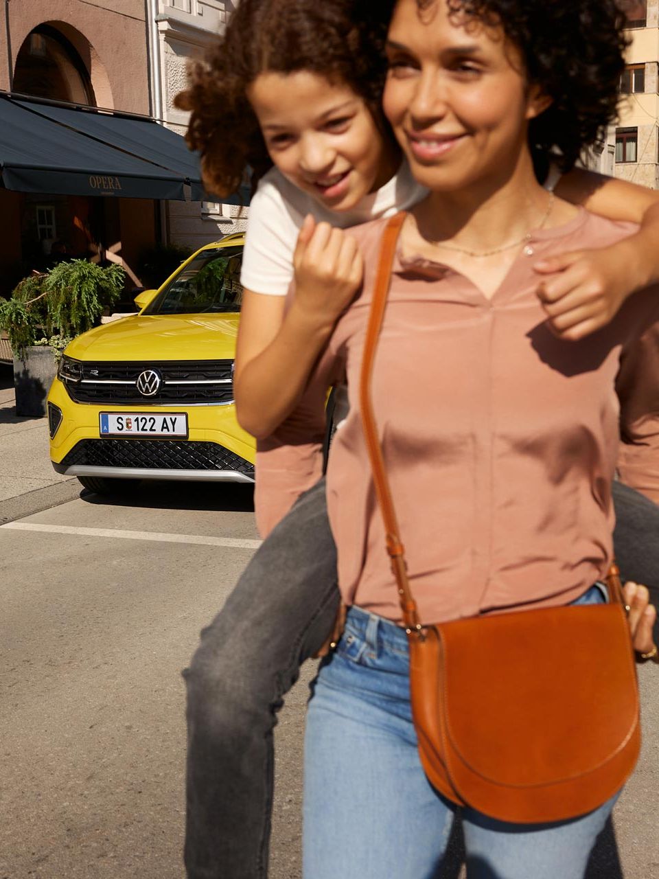 Na ulici v centre mesta nesie žena na chrbte dievča a žltý VW T-Cross stojí v pozadí.