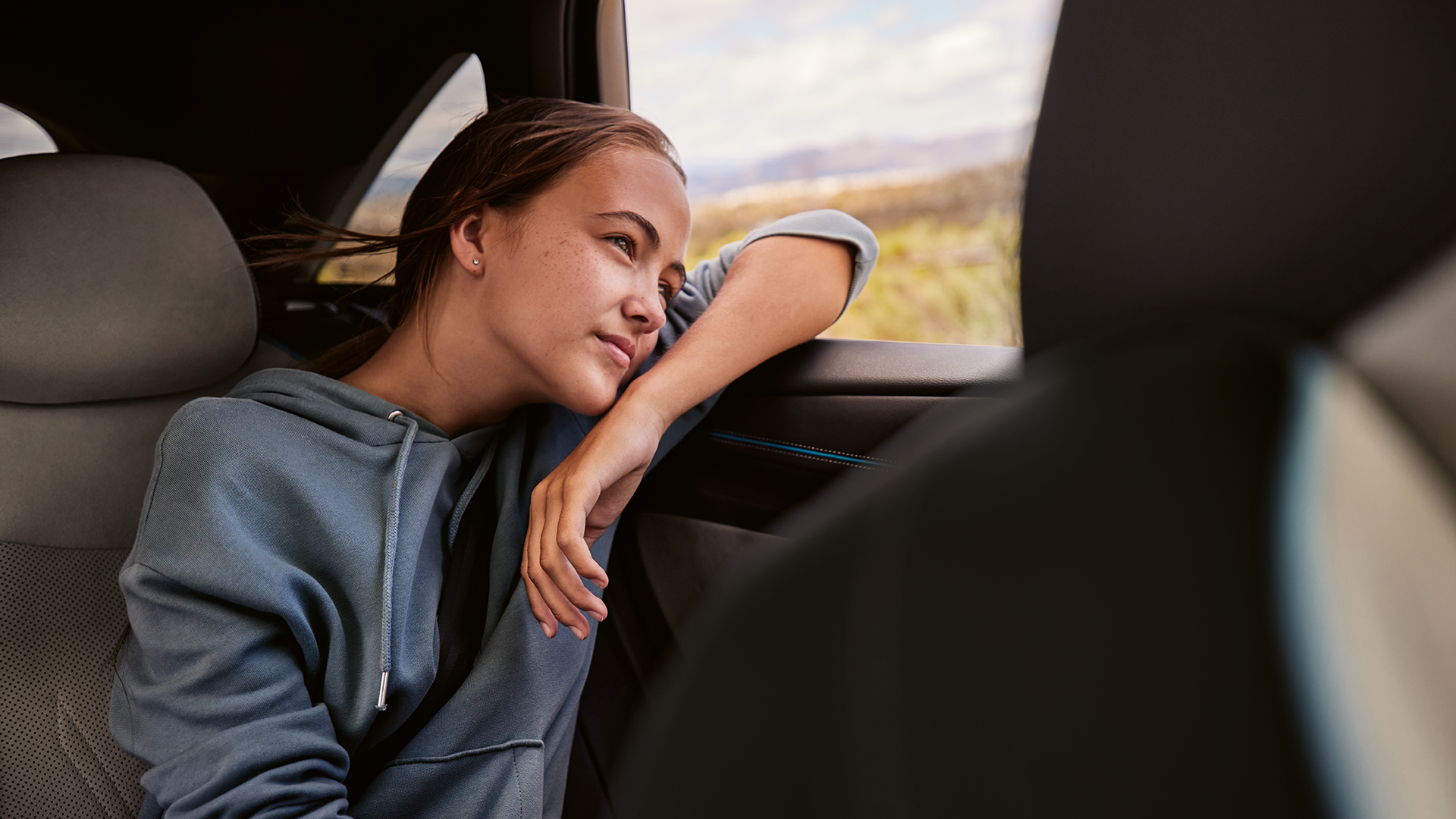 Dievča sedí na zadnom sedadle automobilu, opiera sa o obklad dverí a má stiahnuté okno. Má oblečenú mikinu, za oknom je vidieť prírodu a kopce v pozadí..