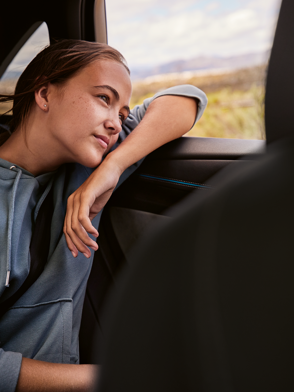 Dievča sedí na zadnom sedadle automobilu, opiera sa o obklad dverí a má stiahnuté okno. Má oblečenú mikinu, za oknom je vidieť prírodu a kopce v pozadí..