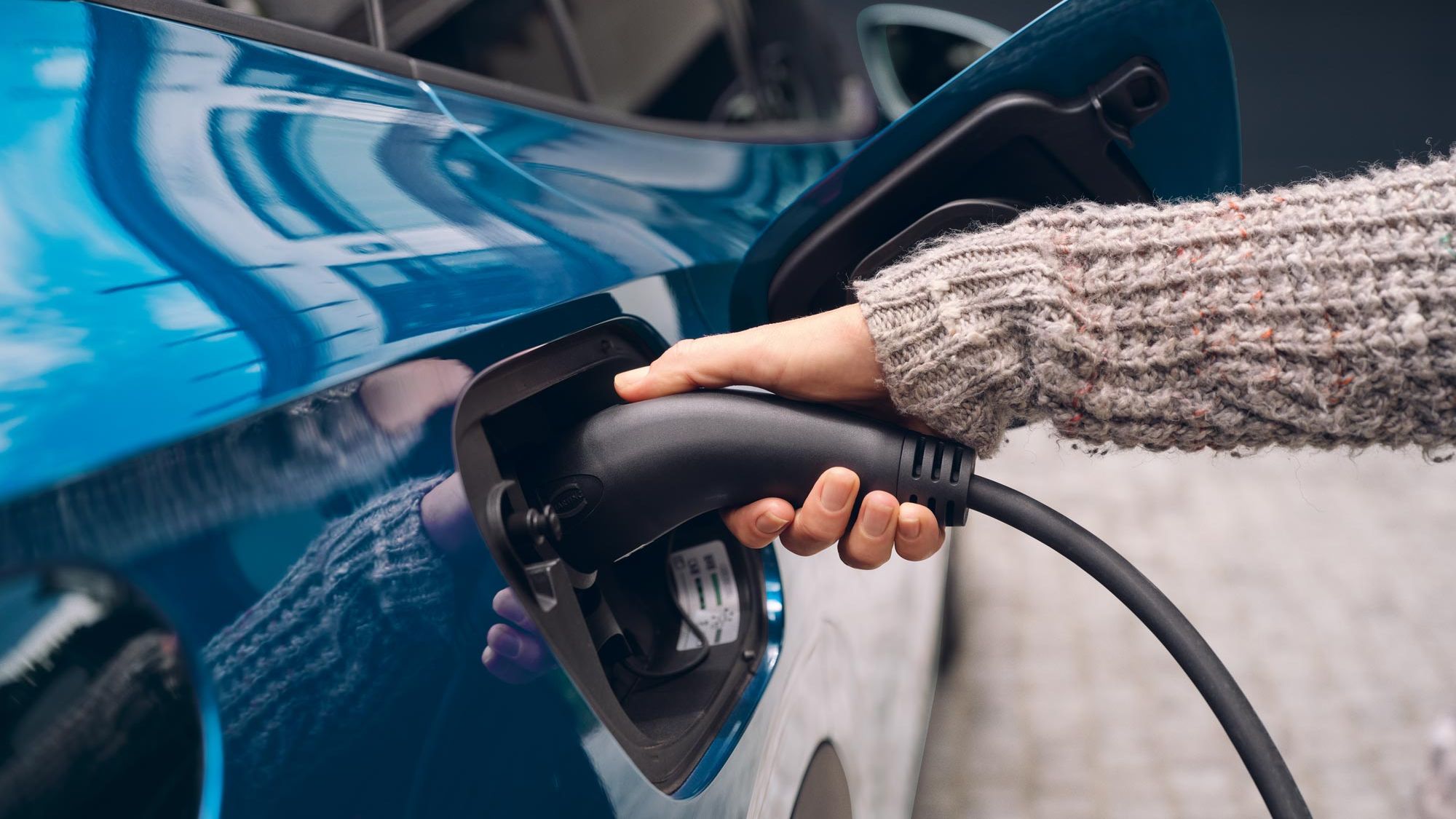 Osoba zapája nabíjací konektor do elektrického auta. Auto je modrej farby.