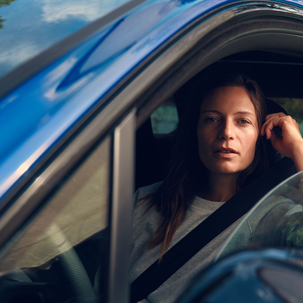 Žena sedí vo vozidle VW Touareg R eHybrid a cez stiahnuté bočné okno sa pozerá priamo do kamery. Za vozidlom je vidieť stromy.