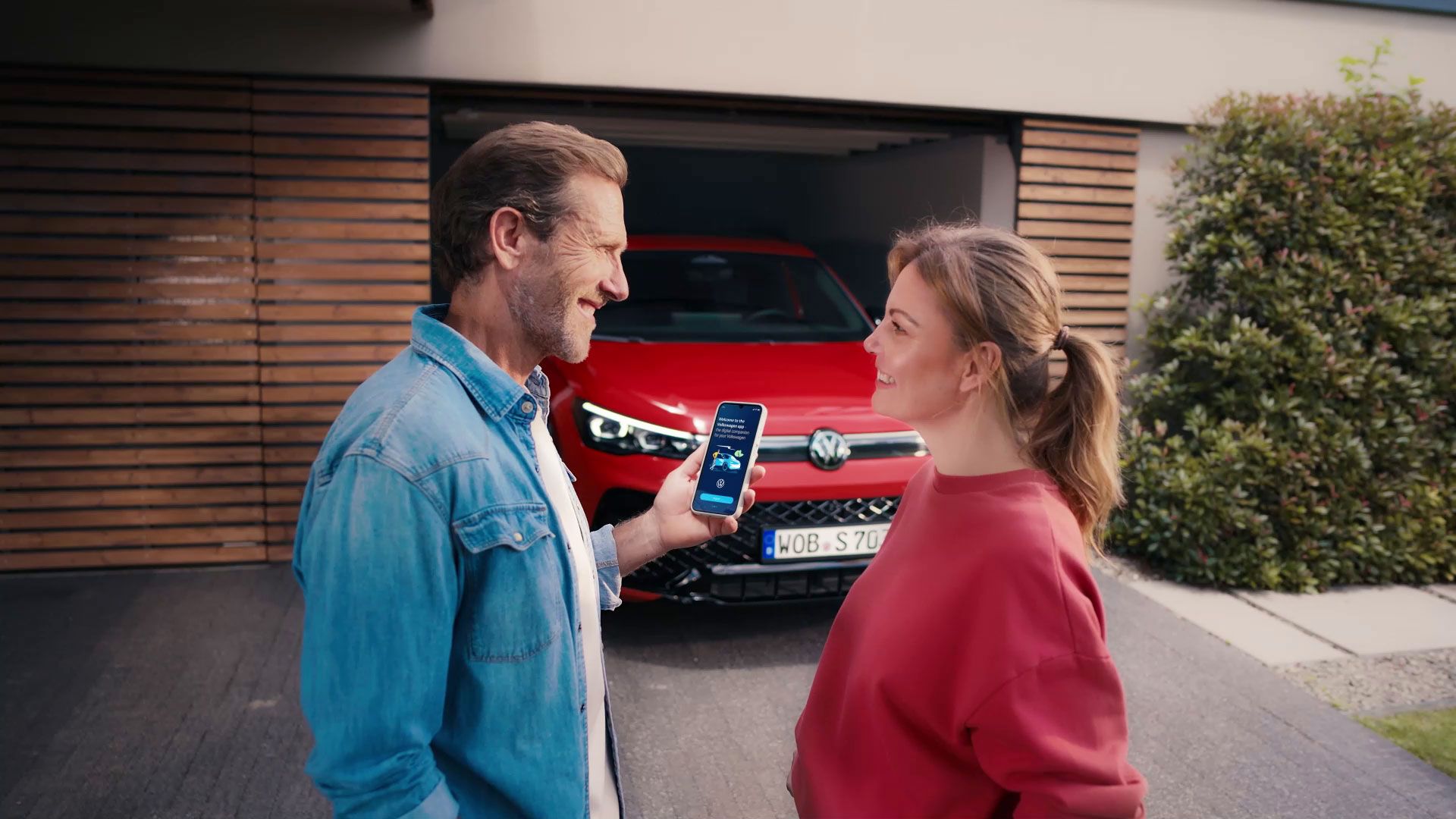 Muž a žena stoja pred garážou, v ktorej je zaparkované červené auto (VW). Muž ukazuje žene niečo na svojom smartfóne. Obe postavy majú rozmazané tváre.