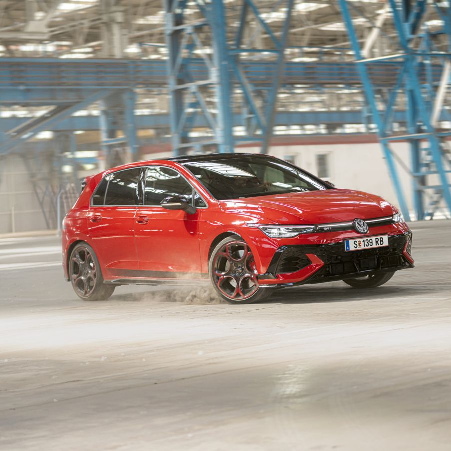 Červený Volkswagen Golf GTI sa preháňa prázdnou priemyselnou budovou. Pneumatiky víria prach. V pozadí sú viditeľné modré oceľové konštrukcie a okná.