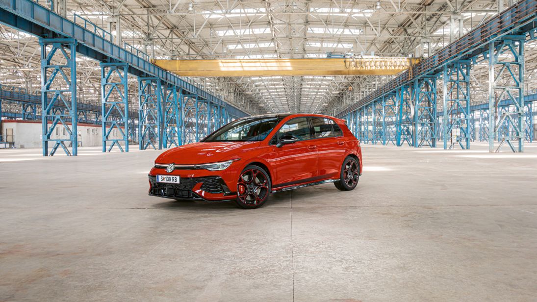 Červený Golf GTI je zaparkovaný vo veľkej, prázdnej priemyselnej budove s modrými oceľovými stĺpmi a nosníkmi. Strecha budovy je zo skla a prepúšťa dovnútra dostatok svetla.