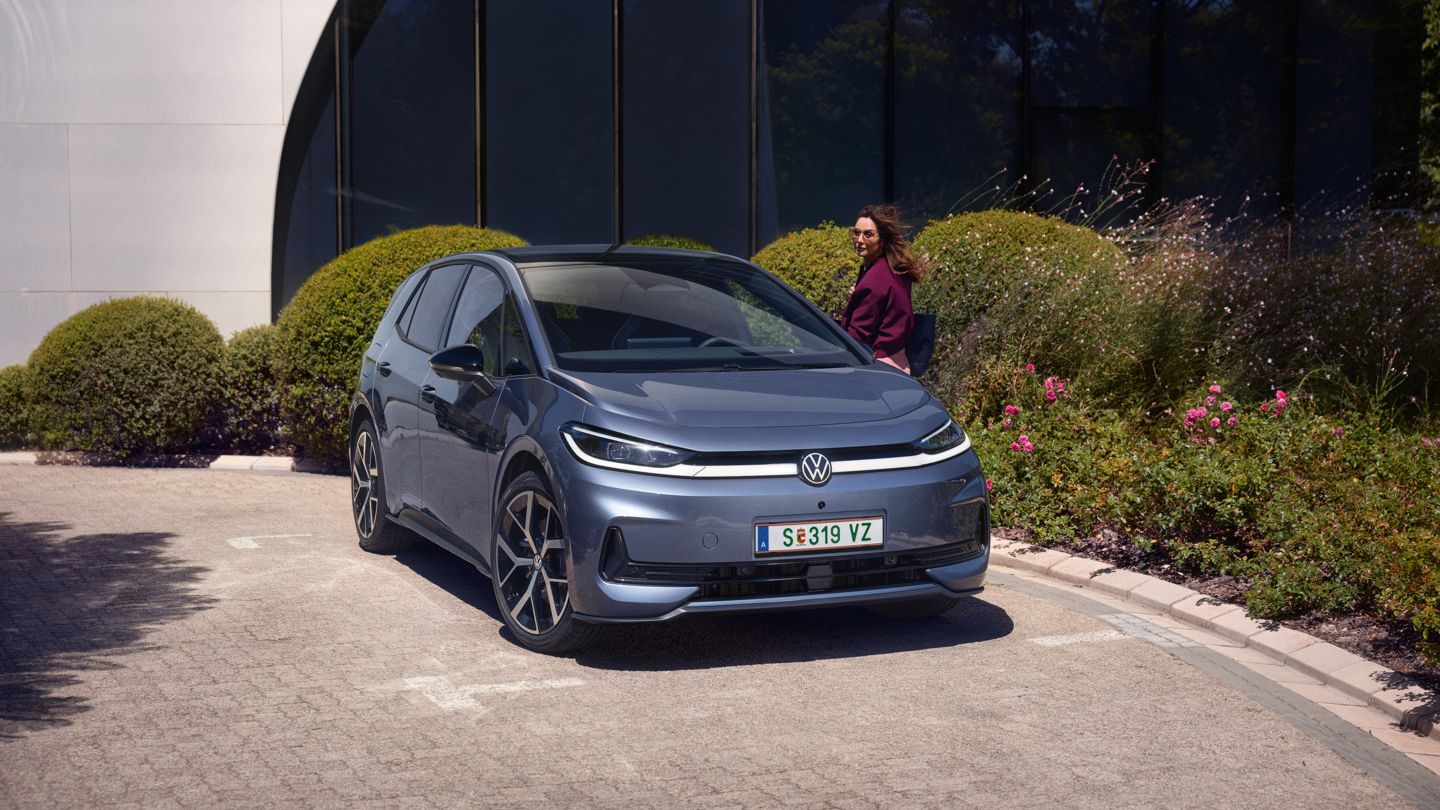 Žena stojí pri dverách vodiča modrého VW ID.3 Neo, ktorý parkuje pred modernou administratívnou budovou.