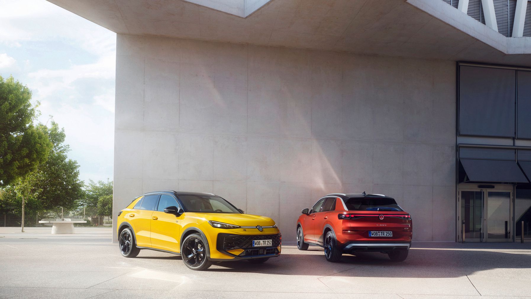 Jeden žltý a jeden červený Volkswagen T-Roc stoja pred vchodom modernej budovy. V pozadí vidieť malé námestie, stromy a park.