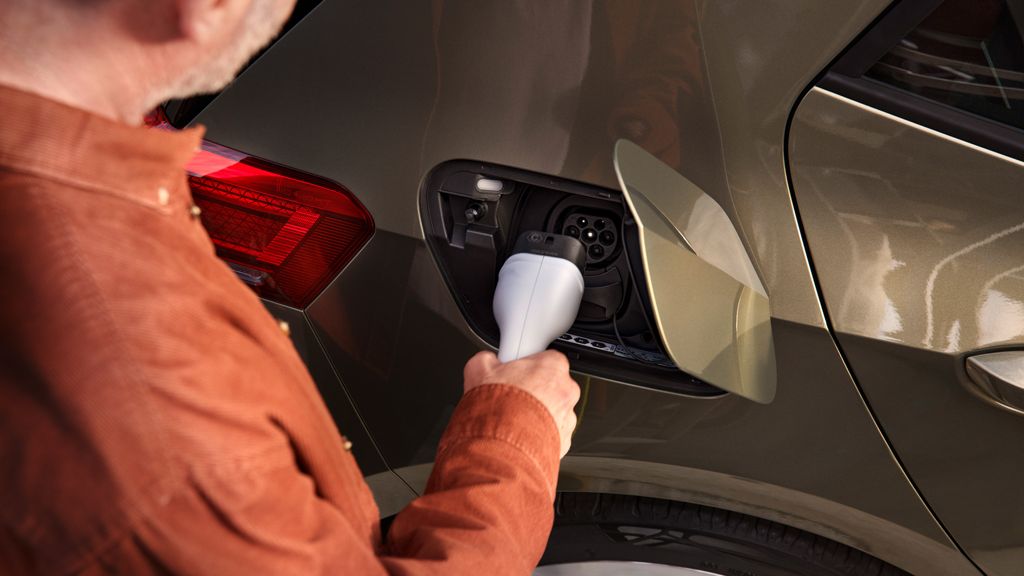 Muž v oranžovom saku nabíja elektrické auto. Nabíjací konektor drží v ruke a vkladá ho do nabíjacieho portu na aute. Auto je olivovo zelené s červeným zadným svetlom.