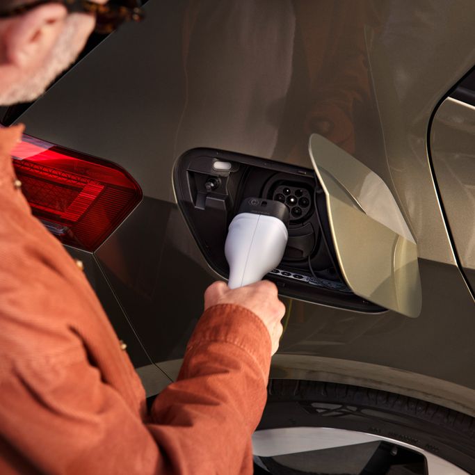 Muž v oranžovom saku nabíja elektrické auto. Nabíjací konektor drží v ruke a vkladá ho do nabíjacieho portu na aute. Auto je olivovo zelené s červeným zadným svetlom.