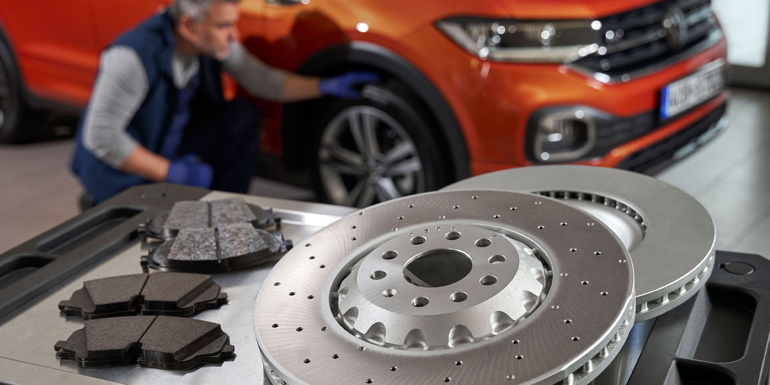 Mechanik kontroluje oranžové SUV, zatiaľ čo na stolíku sú umiestnené nové brzdové kotúče a brzdové doštičky.