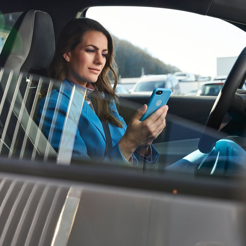 Žena oblečená v modrom sedí v zaparkovanom vozidle Volkswagen so smartfónom v ruke.