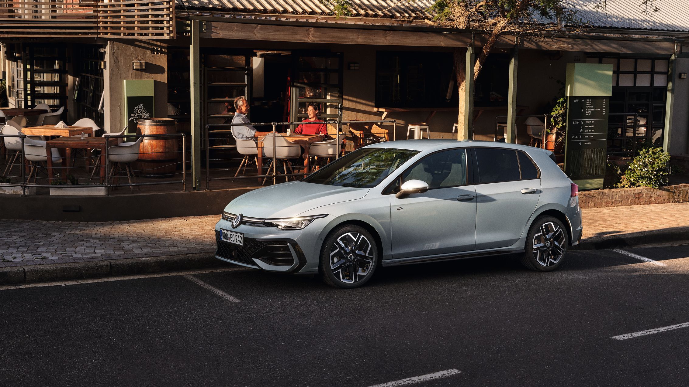 Biely Golf R-Line zaparkovaný na parkovacom mieste na ceste pred reštauráciou. Za vozidlom je reštaurácia a na terase sedia zákazníci.