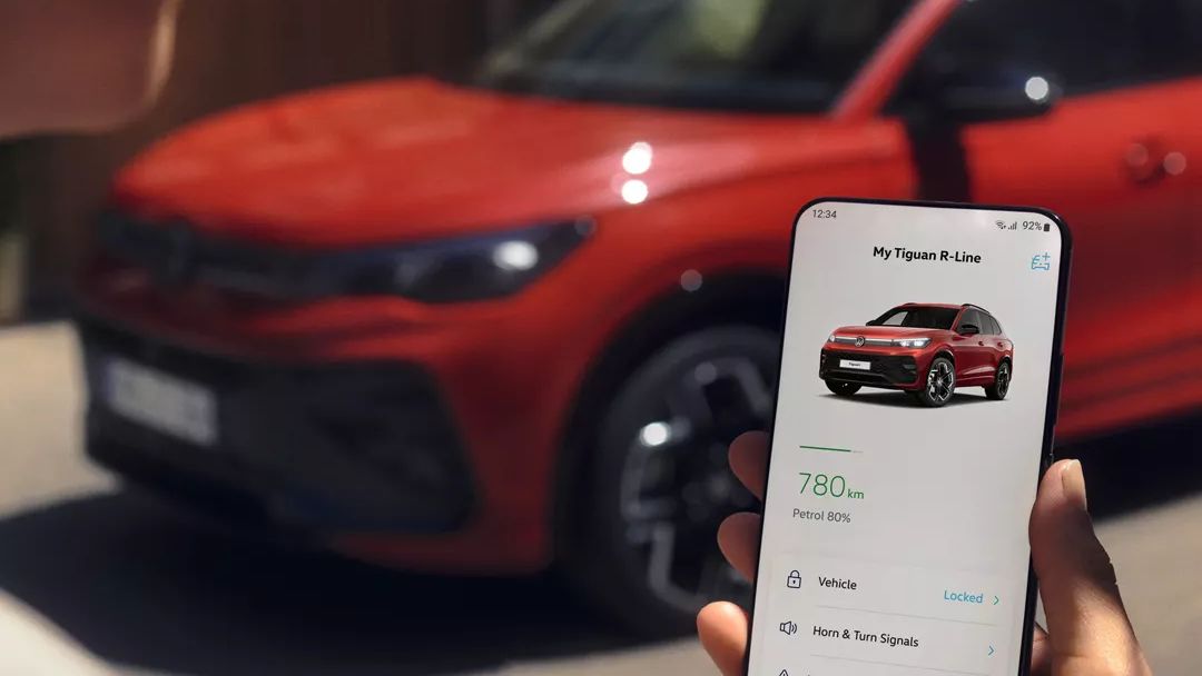 Pohľad cez rameno osoby na mobilný telefón, na ktorom je otvorená aplikácia Volkswagen App. V pozadí stojí červený Tiguan R-Line.