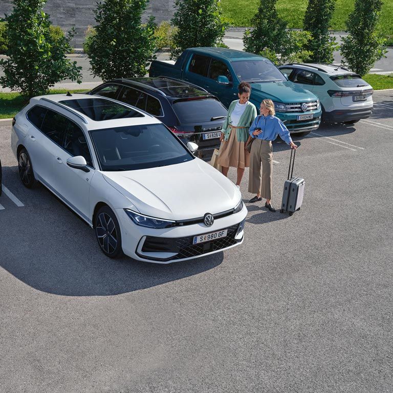 Pohľad mierne zhora na parkujúci biely VW Passat. Pri vozidle stoja dve ženy, jedna drží kufor, druhá tašku.