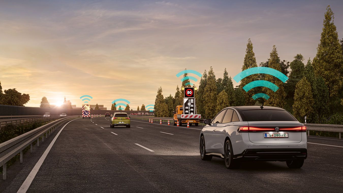 VW Varovanie pred nebezpečenstvom Car2X zobrazené ako modrý ikona WiFi nad vozidlami, ktoré jazdia po diaľnici. Diaľnicu lemuje zeleň a stromy. Atmosféru dotvára modrá, čiastočne zatiahnutá obloha pri západe slnka.