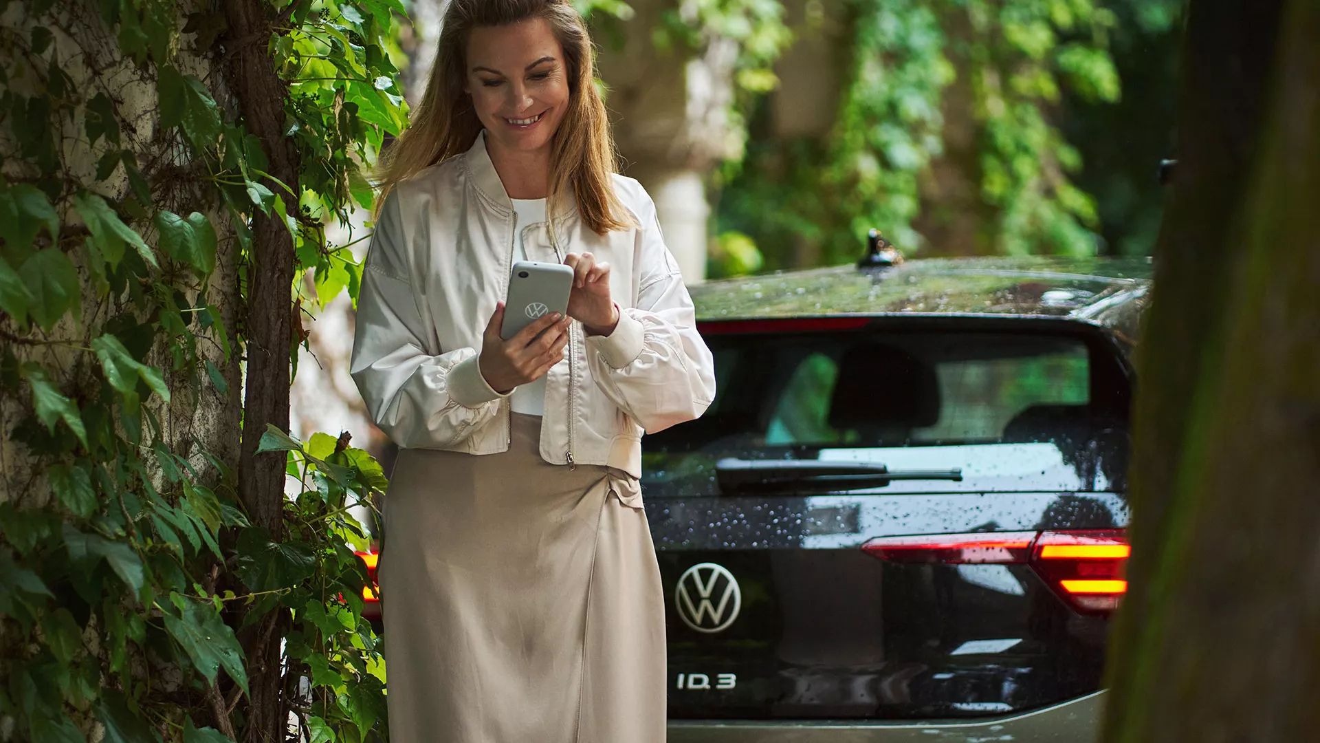 Žena sa pozerá na svoj smartfón a aktivuje režim Battery Care Mode v aplikácii VW, v pozadí je vozidlo ID.3.