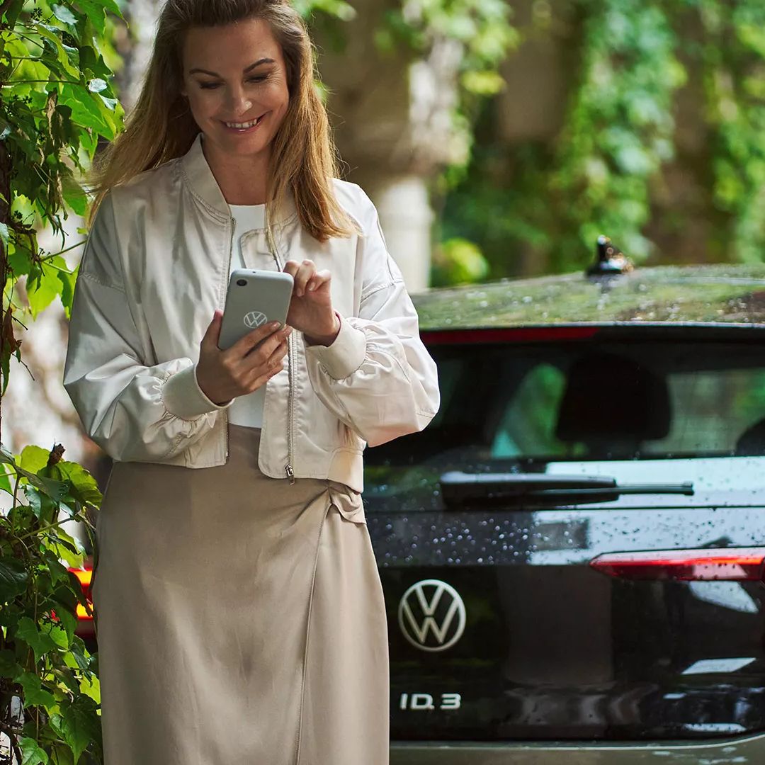 Žena sa pozerá na svoj smartfón a aktivuje režim Battery Care Mode v aplikácii VW, v pozadí je vozidlo ID.3.